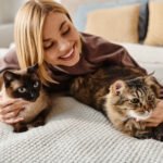 A woman with short hair peacefully lays on a bed, surrounded by two cats, enjoying a moment of tranquility at home.
