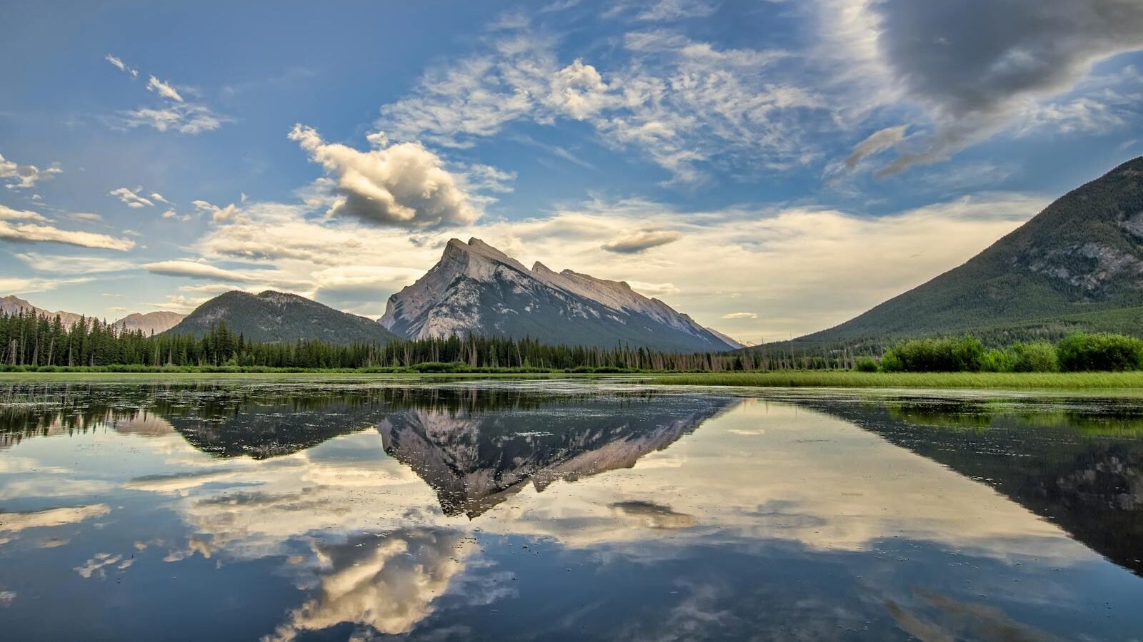 A serene landscape featuring a calm water body reflecting a majestic mountain range under a vibrant sky with scattered clouds. The lush greenery around the water enhances the natural beauty.