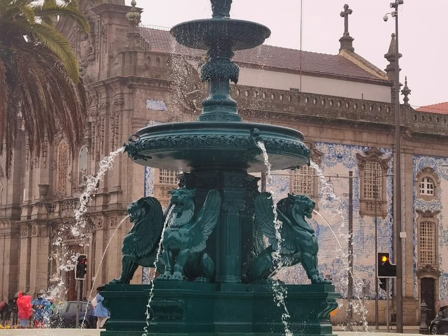 A decorative fountain with lion sculptures spouting water, set against a historical building and palm trees in a public square. 