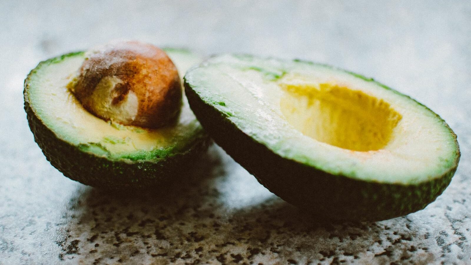 A ripe avocado cut in half, with one half showing the seed and the other the hollowed-out pit area. The avocado is placed on a textured surface, highlighting its vibrant green interior and rough dark skin.