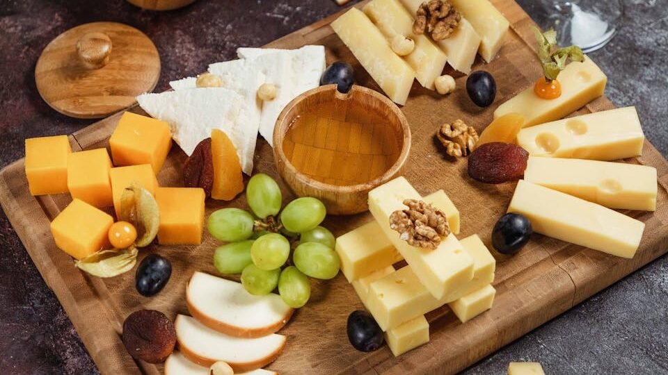 A wooden cheese board arranged with a variety of cheeses, fresh green grapes, sliced apples, dried fruits, nuts, and a small bowl of honey. The setting includes a glass of white wine, creating a sophisticated and appetizing presentation.