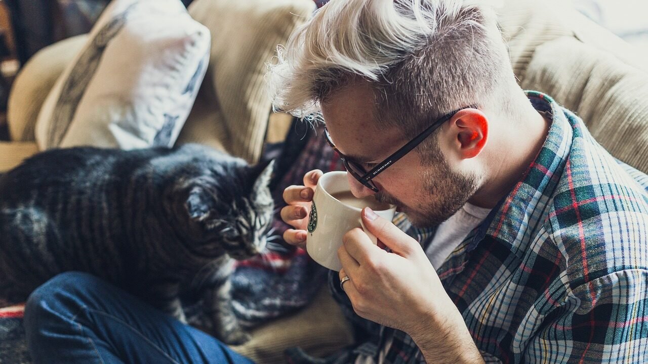 A man sitting on a couch with a cup of coffee, leaning forward as a black cat curiously sniffs the cup