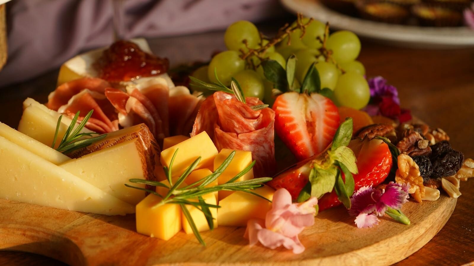 A beautifully arranged charcuterie board featuring an assortment of cheeses, sliced cured meats, fresh strawberries, green grapes, nuts, and garnishes of rosemary and edible flowers.