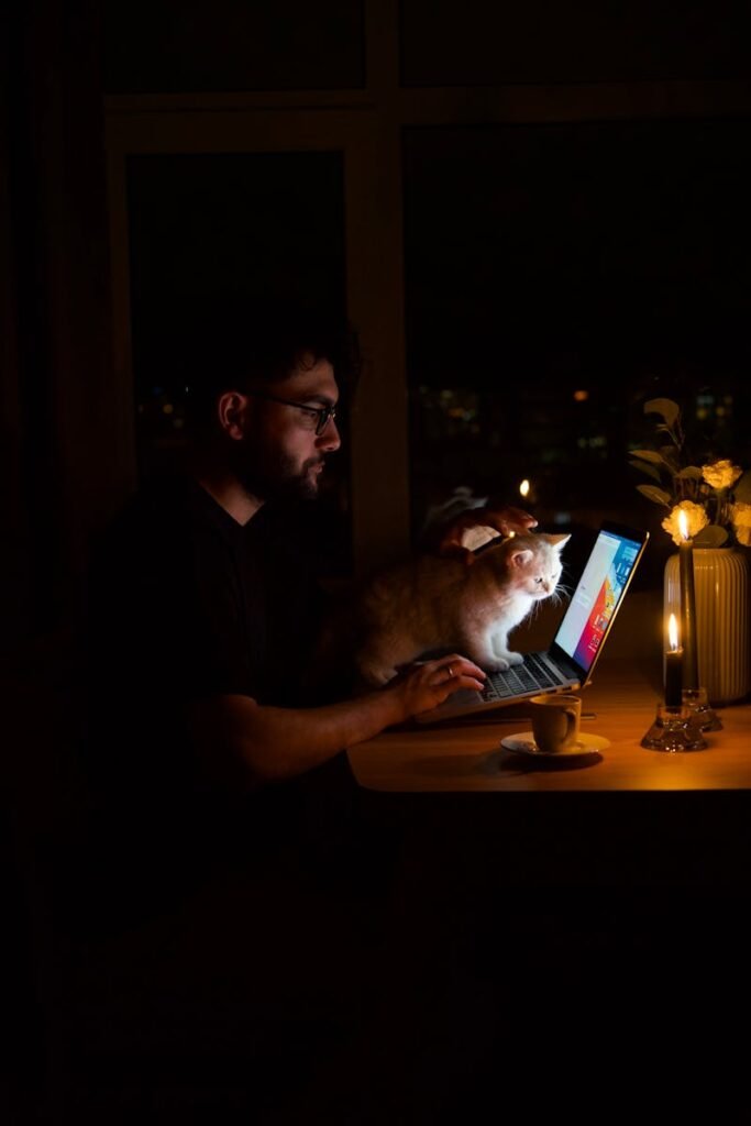 Man using laptop at night with a cat beside him, illuminated by candlelight.