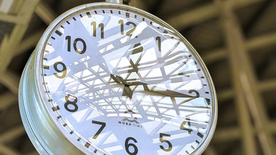 A large clock hanging in a brightly lit space with reflections and shadows cast by an overhead structure. The clock displays a clear time, symbolizing precision and timeliness.