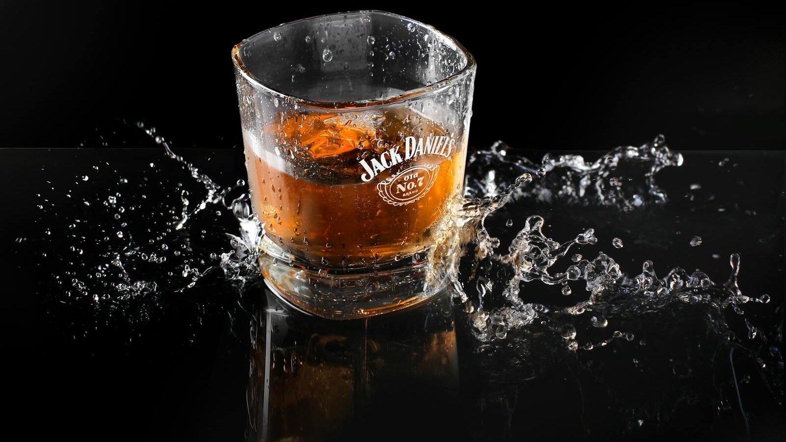 A glass of Jack Daniel's whiskey with ice, sitting on a reflective black surface. Splashing water surrounds the glass, creating a dynamic and dramatic effect.