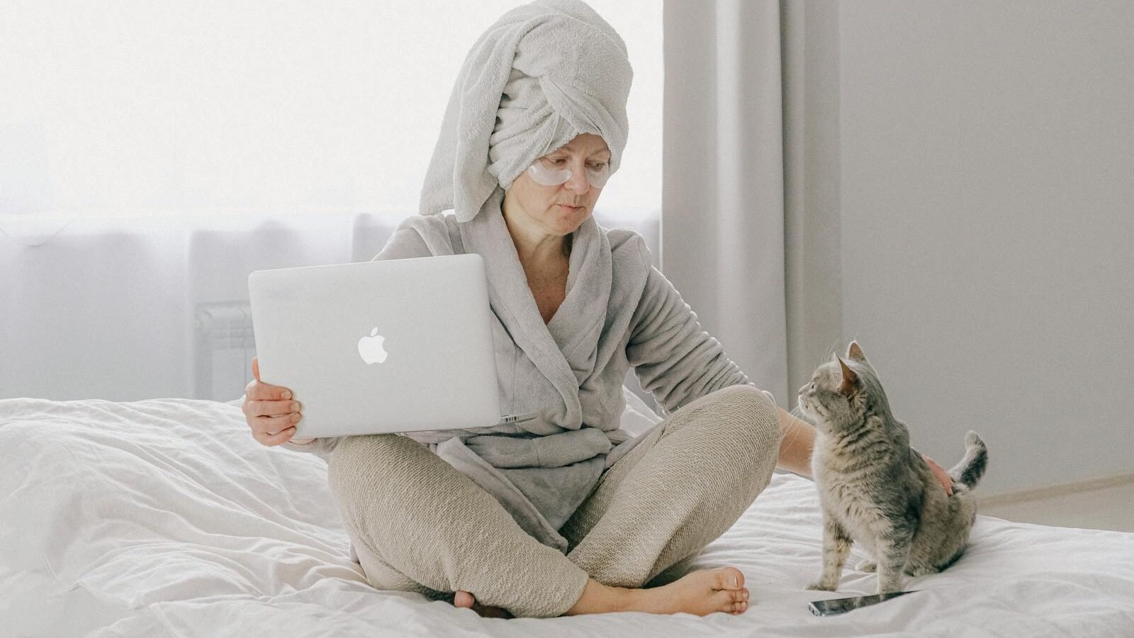 A woman sitting on a bed in a cozy bathrobe with a towel wrapped around her head, holding a laptop. A gray tabby cat sits nearby, looking up at her, showcasing a peaceful interaction in their routine.