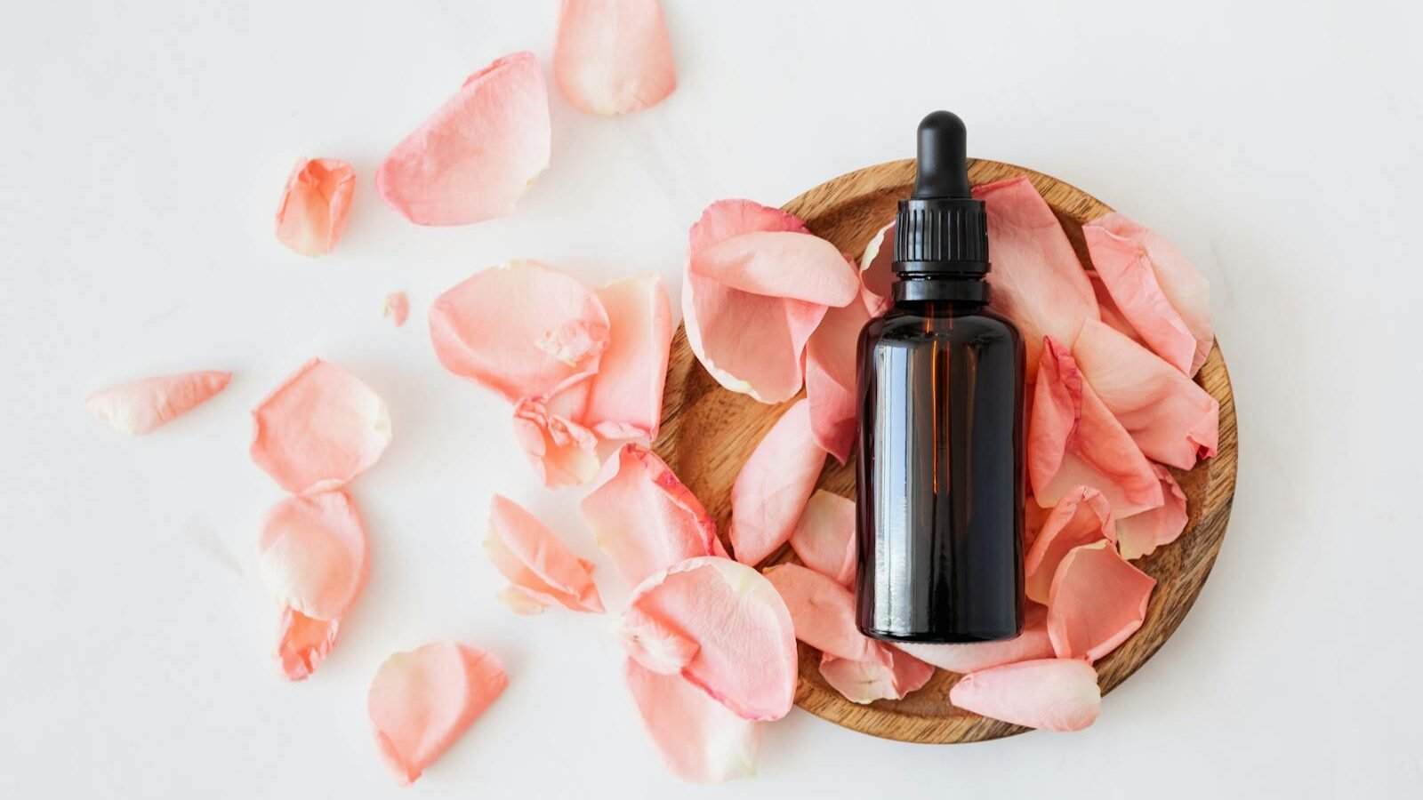 A brown glass dropper bottle placed on a wooden dish surrounded by soft pink rose petals, set against a clean white background, evoking a sense of natural beauty and skincare.