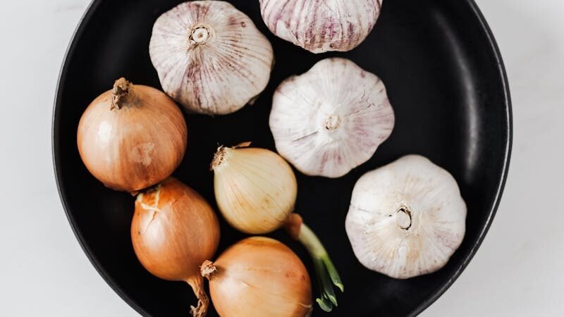 A black plate containing several whole garlic bulbs and onions, arranged neatly on a white background. The contrast between the dark plate and the light-colored ingredients highlights their natural textures.