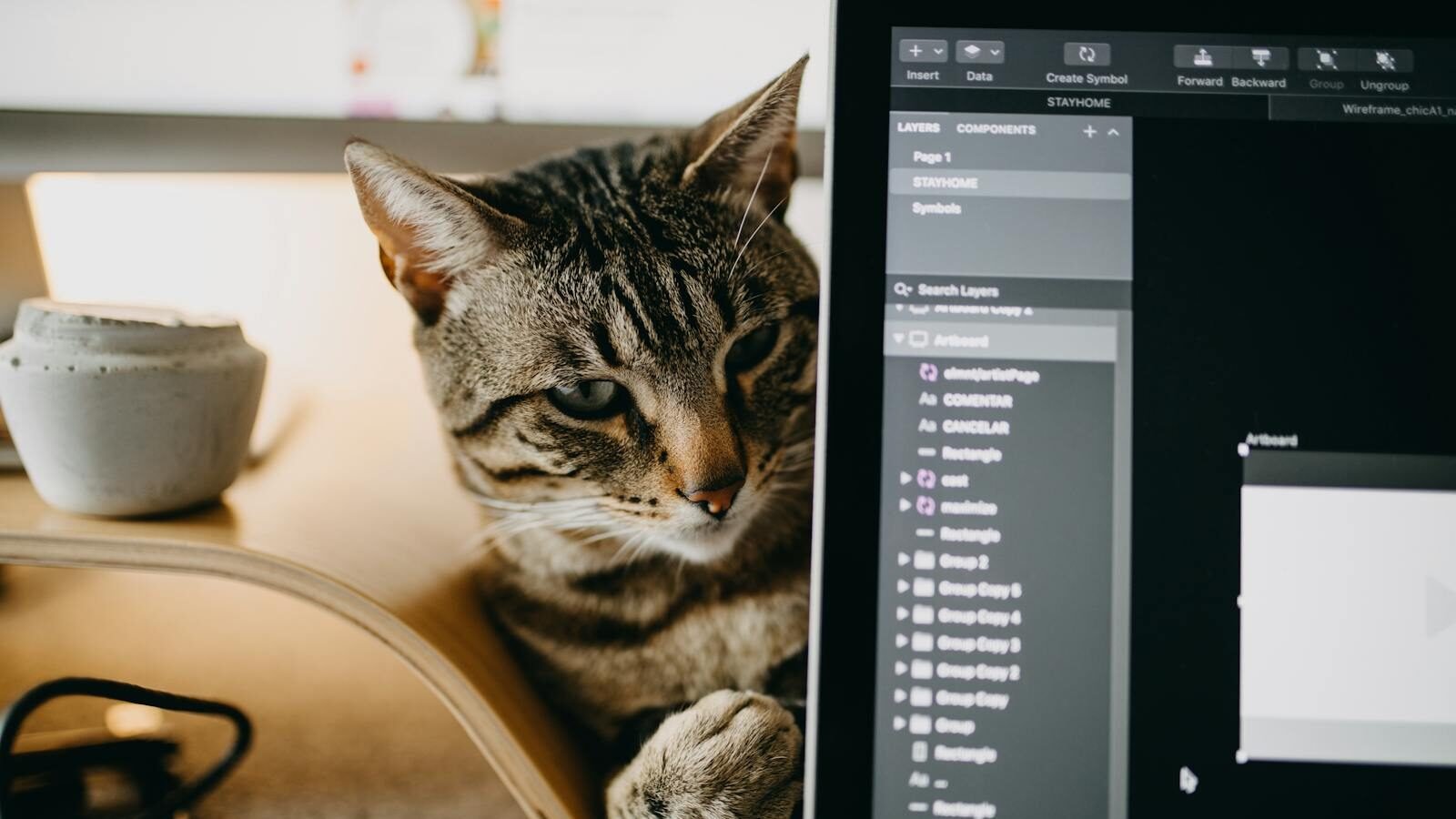 A tabby cat sitting next to a computer screen displaying a file management system, with a coffee mug and desk accessories in the background.