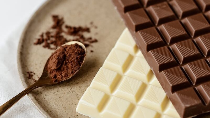 Two chocolate bars, one milk chocolate and one white chocolate, placed on a ceramic plate alongside a spoon of cocoa powder and some scattered cocoa flakes.