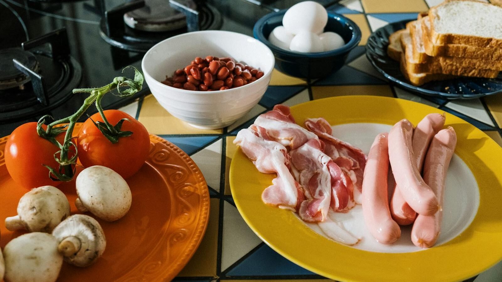 A kitchen counter featuring ingredients for a meal, including raw bacon and sausages on a yellow plate, tomatoes and mushrooms on an orange plate, a bowl of red beans, eggs in a small dish, and slices of bread. The background includes a tiled surface with a gas stove.