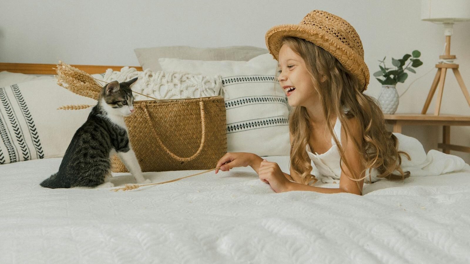 Joyful child in a straw hat playing with a cat on a bed, surrounded by cozy decor and plants.