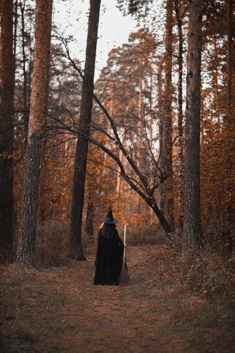 Photo by Monstera Production via Pexels A witch in a black costume walks through an eerie autumn forest, capturing the essence of Halloween.
