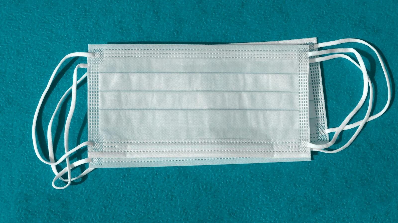 A pair of white disposable face masks placed on a teal surface. The masks are shown flat, highlighting their pleated design and ear loops, symbolizing tools for controlling disease spread.