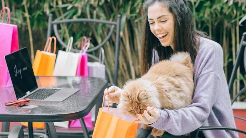 A smiling woman sitting at an outdoor table with shopping bags, a laptop, and a fluffy orange cat on her lap, enjoying a relaxing moment.
