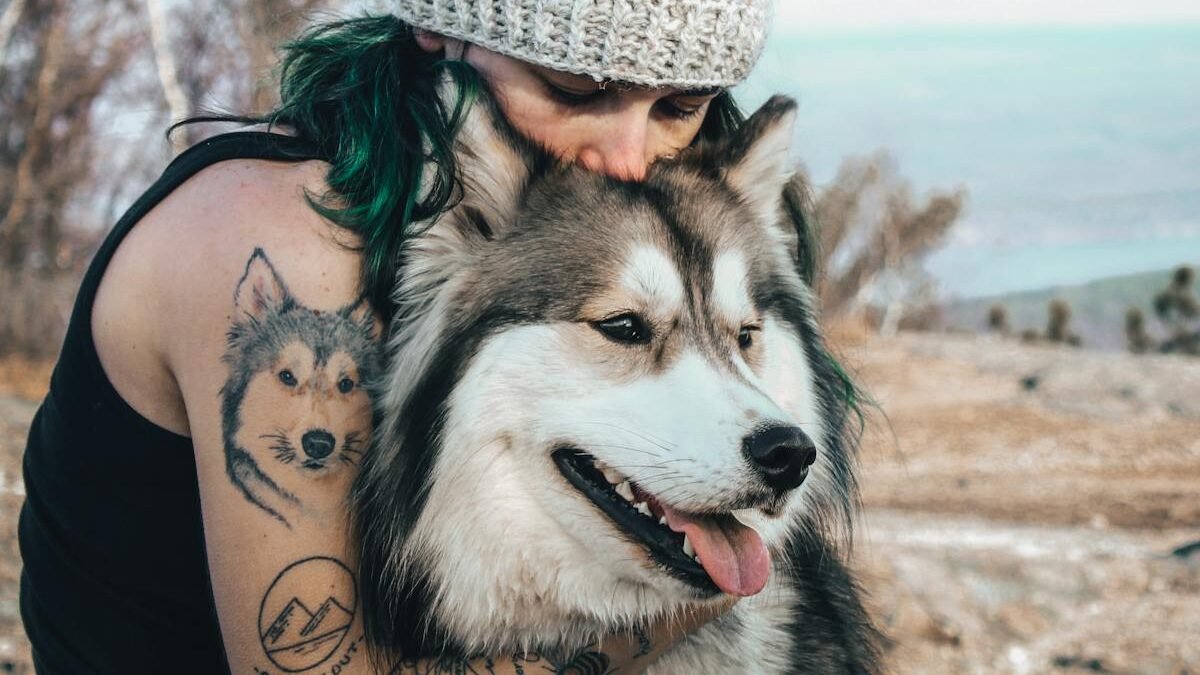 A woman wearing a knit hat and showcasing dog-themed tattoos hugs a Siberian Husky outdoors. The Husky appears happy and relaxed, highlighting the strong bond and affection between humans and dogs.