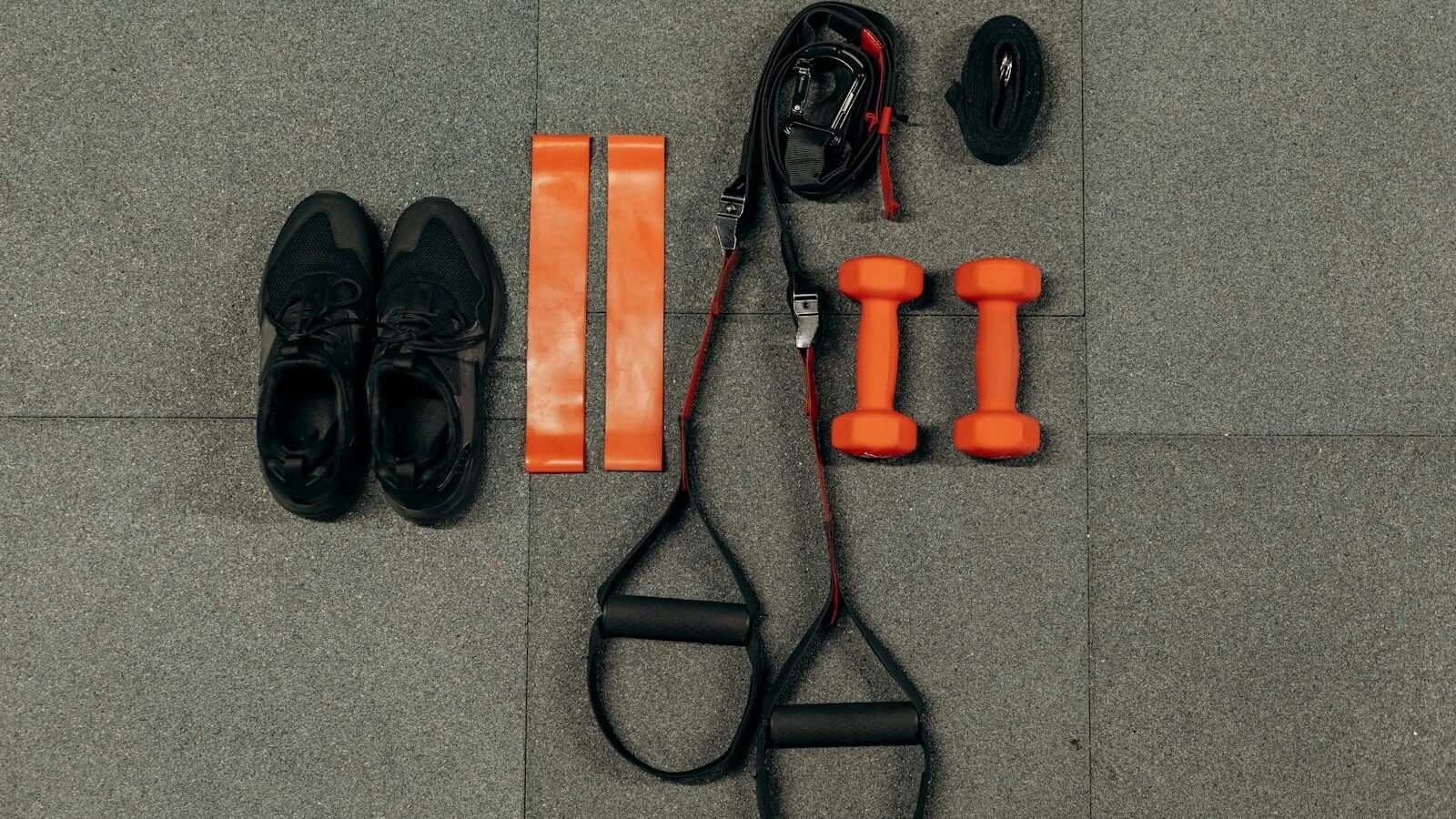 A set of training tools including black shoes, orange resistance bands, orange dumbbells, and black handles arranged neatly on a gray tiled floor.