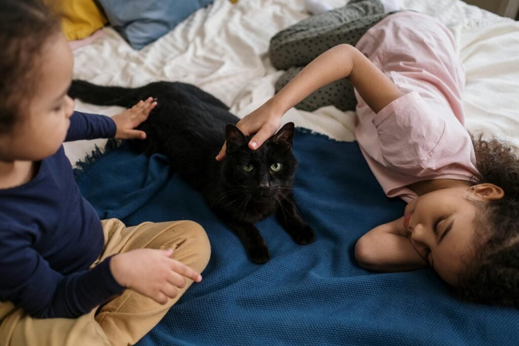 Two children relaxing on a bed with a black cat, one petting its head while the other watches, creating a calm and loving atmosphere