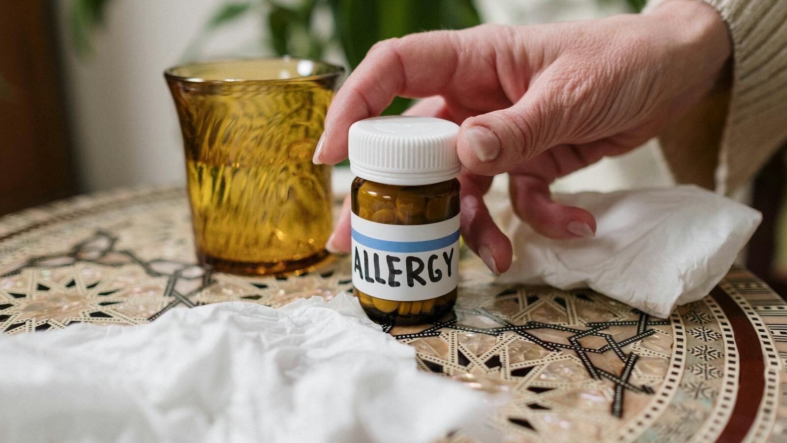 A close-up of a hand reaching for a small bottle labeled "Allergy" on a decorative table, surrounded by tissues and a glass.