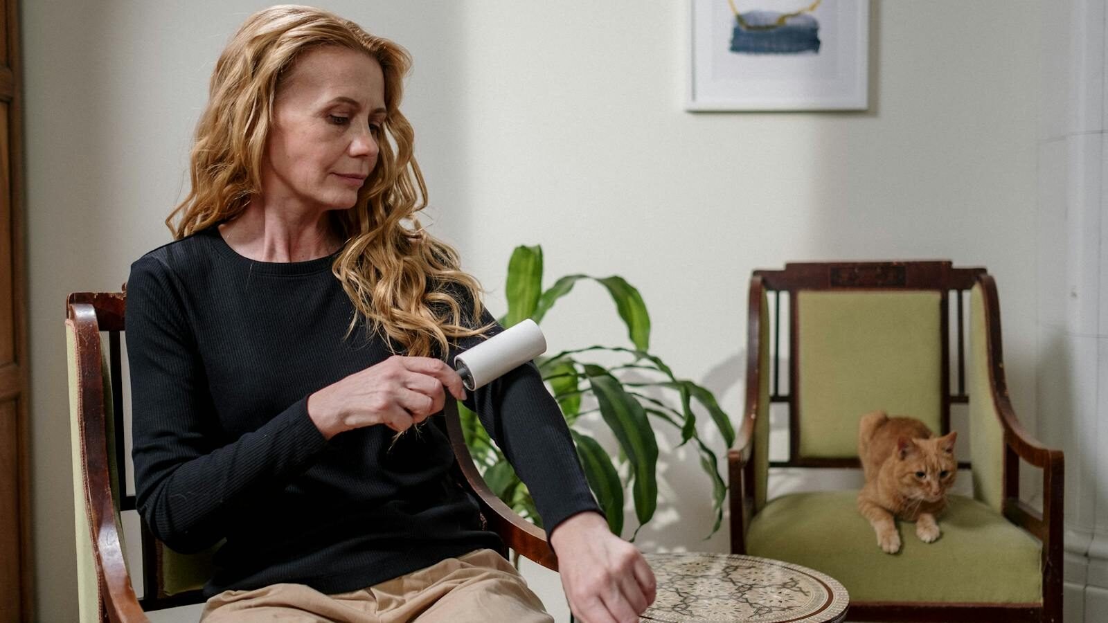 A woman using a lint roller on her black sweater while sitting in a living room, with an orange cat lounging on a chair in the background.