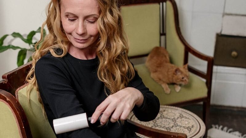 A woman with long blonde hair using a lint roller on her black shirt while sitting on a green chair, with an orange cat lounging on a chair in the background.