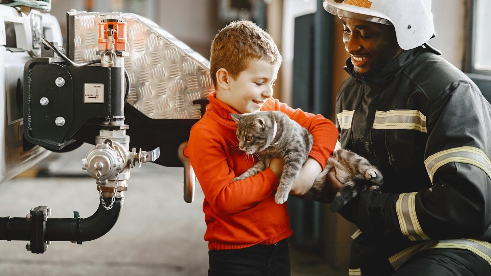 A firefighter wearing protective gear smiles while helping a young boy hold a gray tabby cat. The scene, set near firefighting equipment, highlights professional assistance in safely handling and rescuing cats.