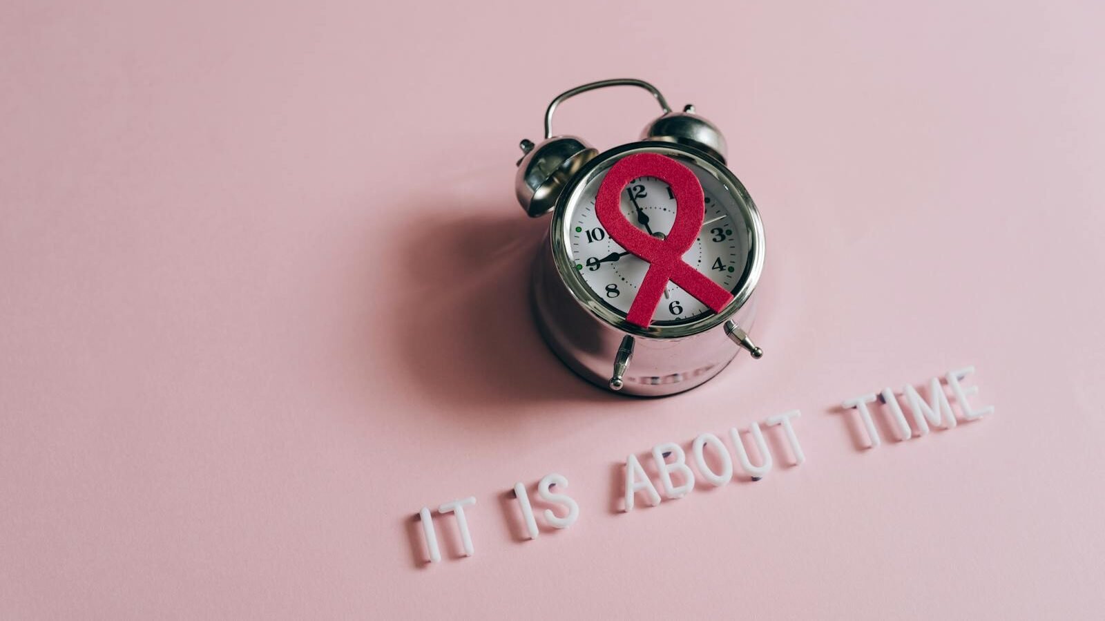 A vintage alarm clock with a pink ribbon symbol on its face, placed on a pink background with the words "IT IS ABOUT TIME" arranged below it, symbolizing awareness and the right time for action.