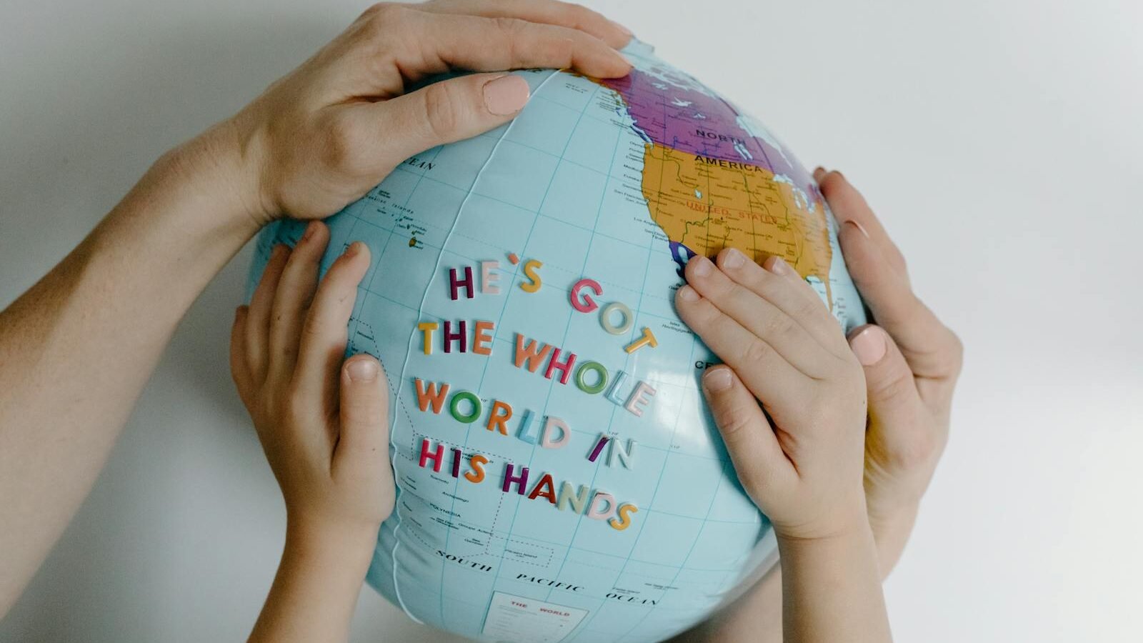 Multiple hands of different sizes holding a globe with the words "He’s got the whole world in his hands" on it.