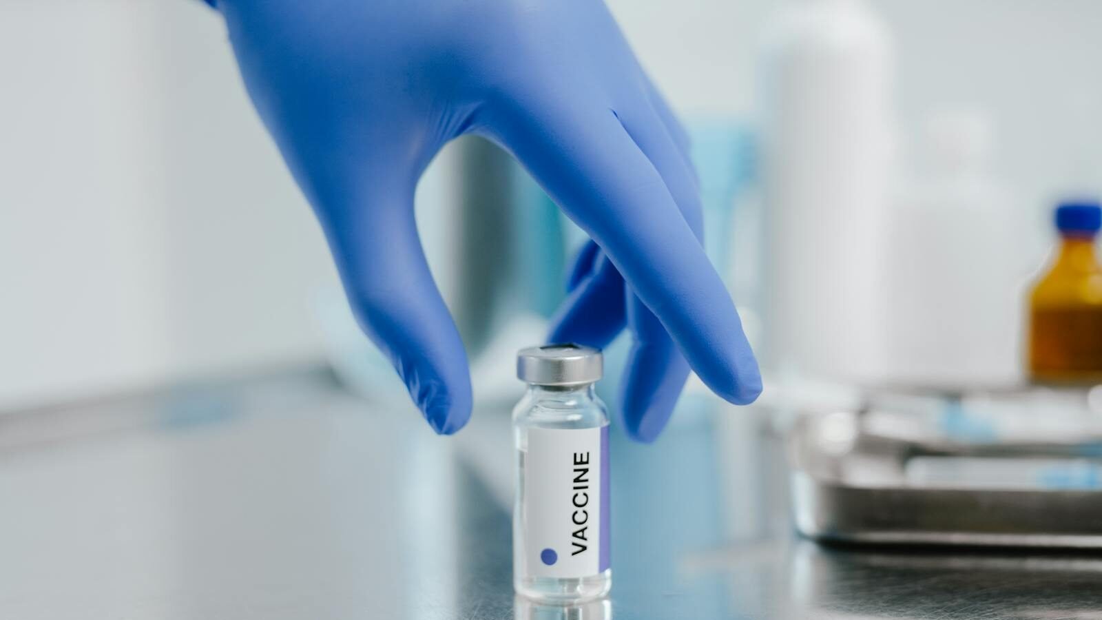 A gloved hand holding a vial labeled "Vaccine" on a reflective medical table, representing allergen immunotherapy treatment.