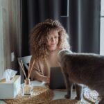 A woman with curly hair sitting at a table, smiling at a gray cat standing on the table. The scene depicts a cozy and friendly interaction, highlighting the bond between humans and cats.