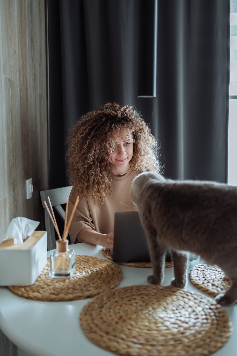 A woman with curly hair sitting at a table, smiling at a gray cat standing on the table. The scene depicts a cozy and friendly interaction, highlighting the bond between humans and cats.