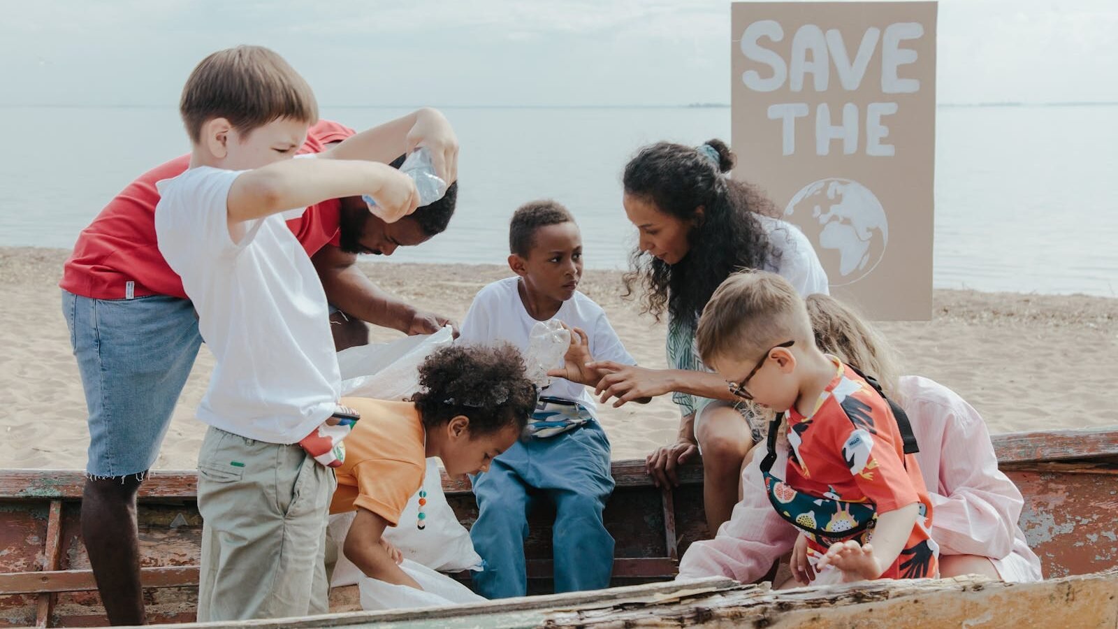 A group of children and a teacher engaging in an outdoor environmental activity near the beach, with a "Save the Earth" sign in the background.