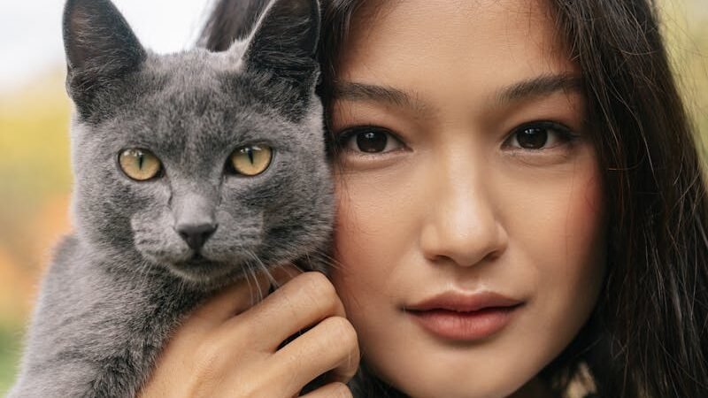 A woman holding a gray cat close to her face, both gazing directly at the camera, highlighting their close bond and connection.