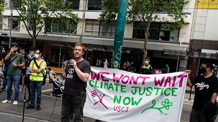 A protester speaking at a public demonstration, with a banner behind them advocating for climate justice and wildlife protection.