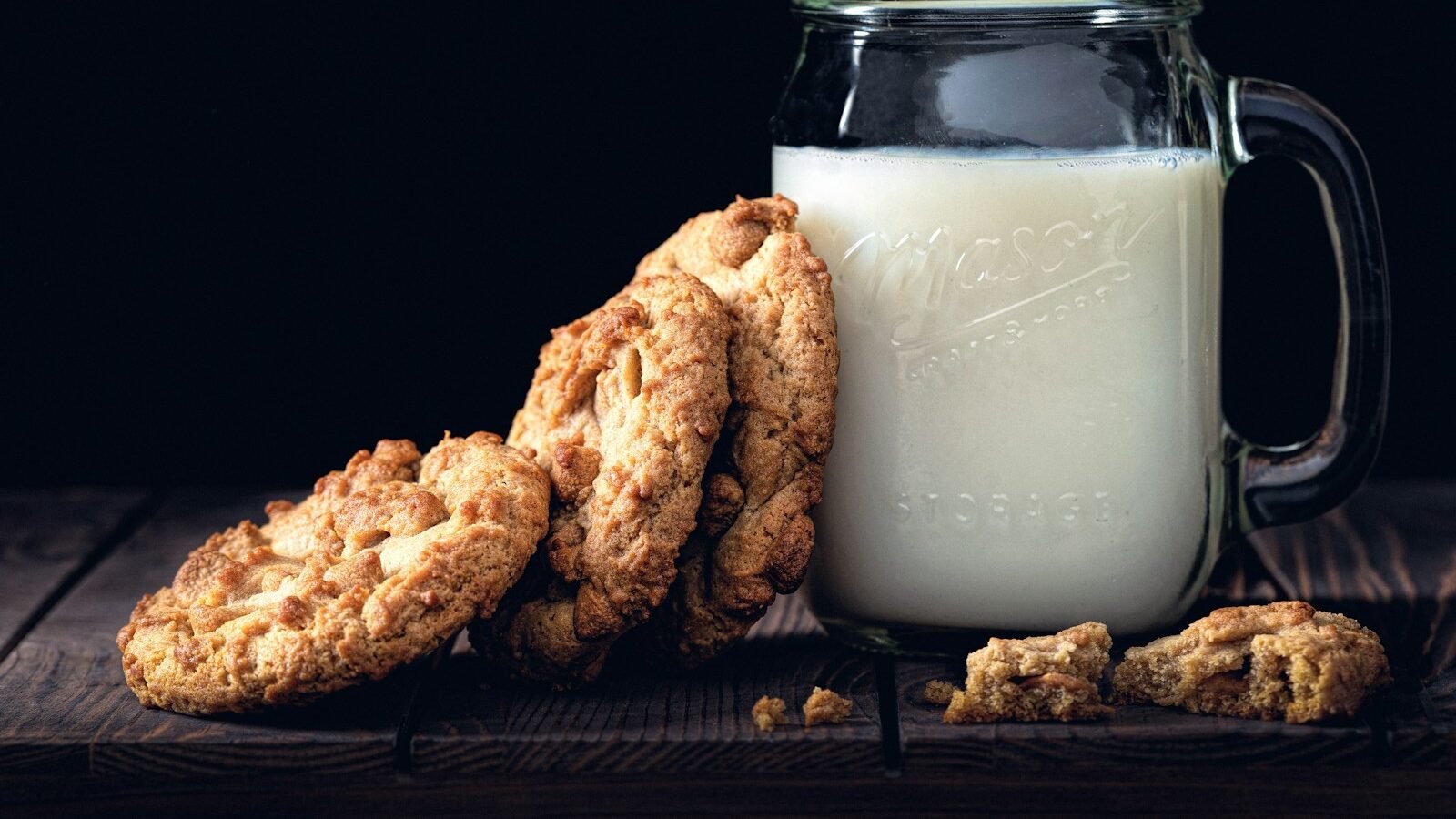 A glass mason jar filled with milk, accompanied by three freshly baked cookies leaning against it. Crumbs are scattered on a rustic wooden table, set against a dark background.