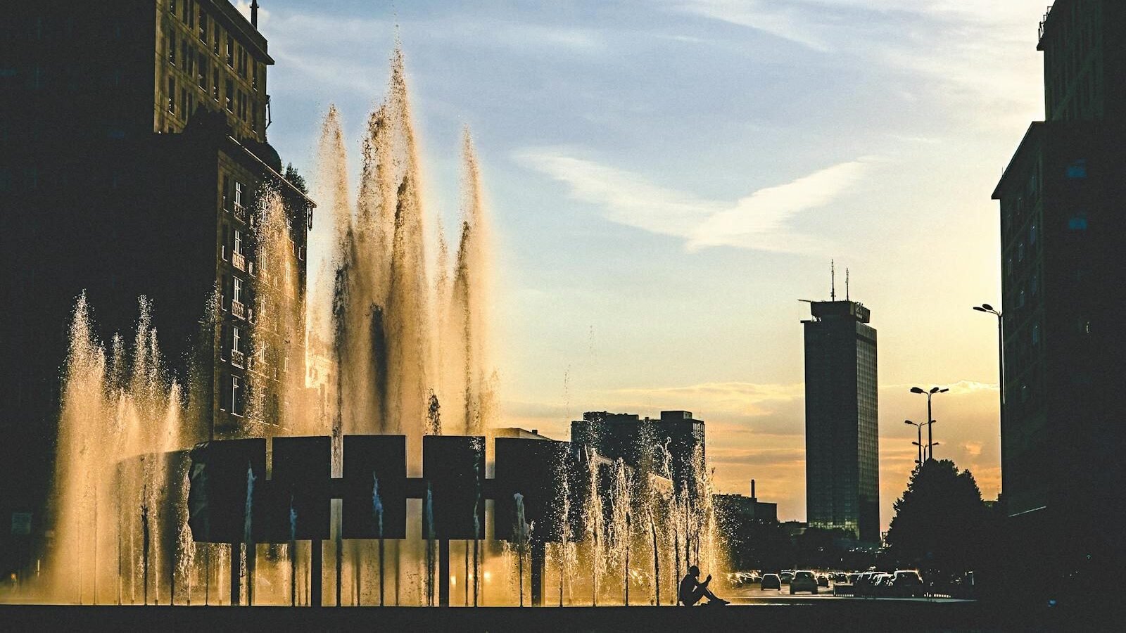 A modern urban scene featuring tall buildings and a beautifully lit water fountain with jets rising high against the backdrop of a sunset sky.