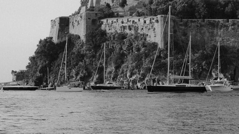 A black-and-white image of an ancient coastal fortress perched on a rocky hill, with sailboats anchored in the water below, evoking the essence of early maritime history.