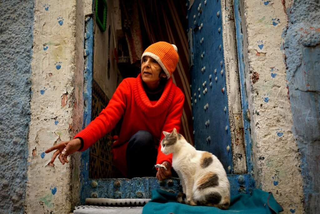 Elderly woman in orange beanie kneeling in the doorway with a cat in Casablanca, Morocco.