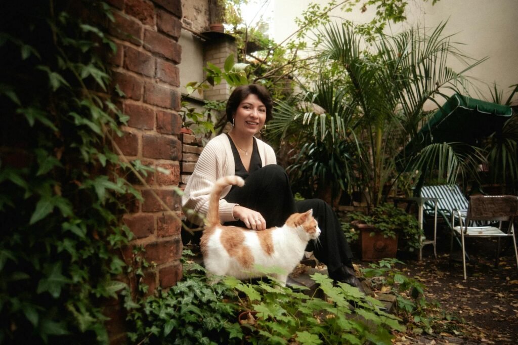 A woman sits in a lush Berlin backyard garden, petting a cat, exuding warmth and relaxation.