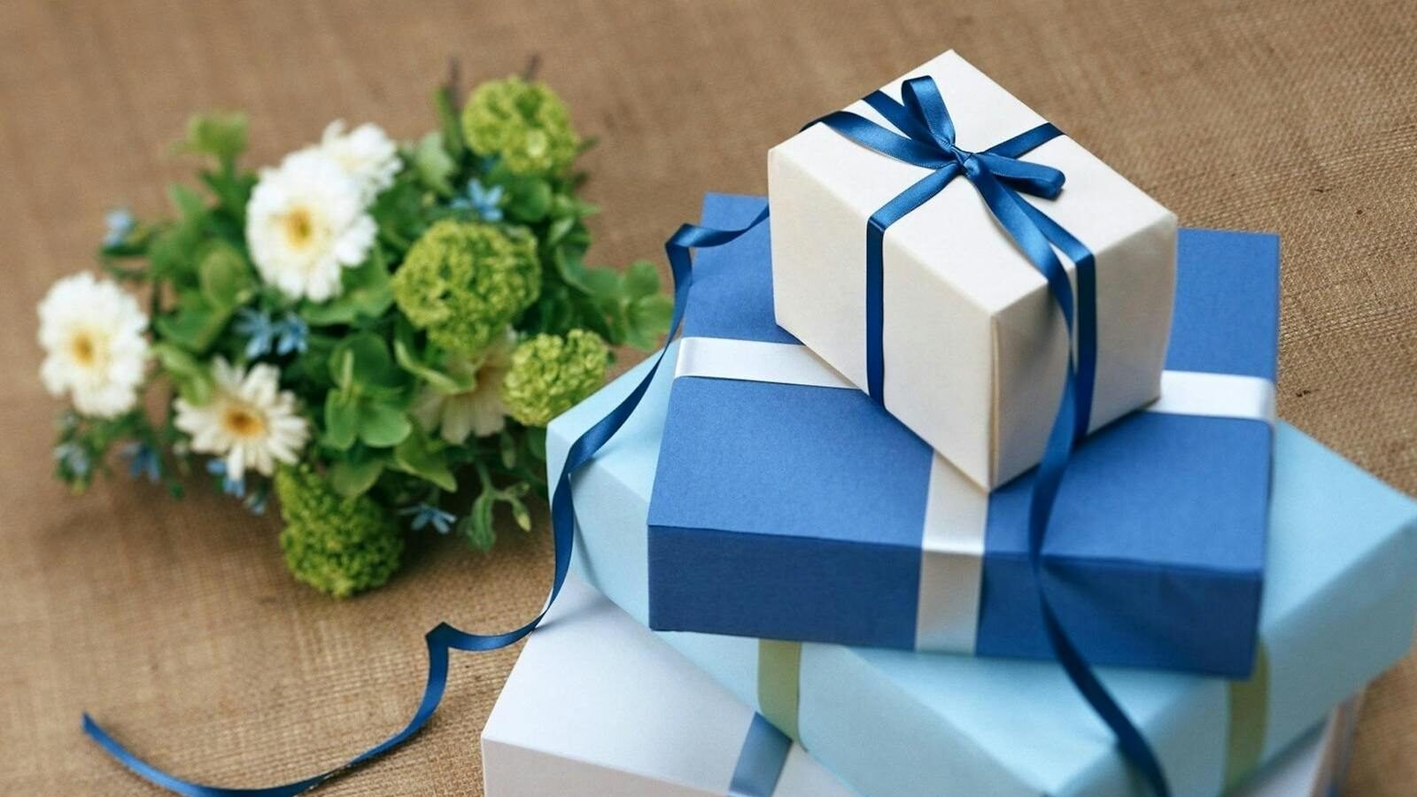 A neatly arranged stack of three gift boxes wrapped in blue and white paper with matching ribbons, placed on a burlap surface next to a small bouquet of white and green flowers.