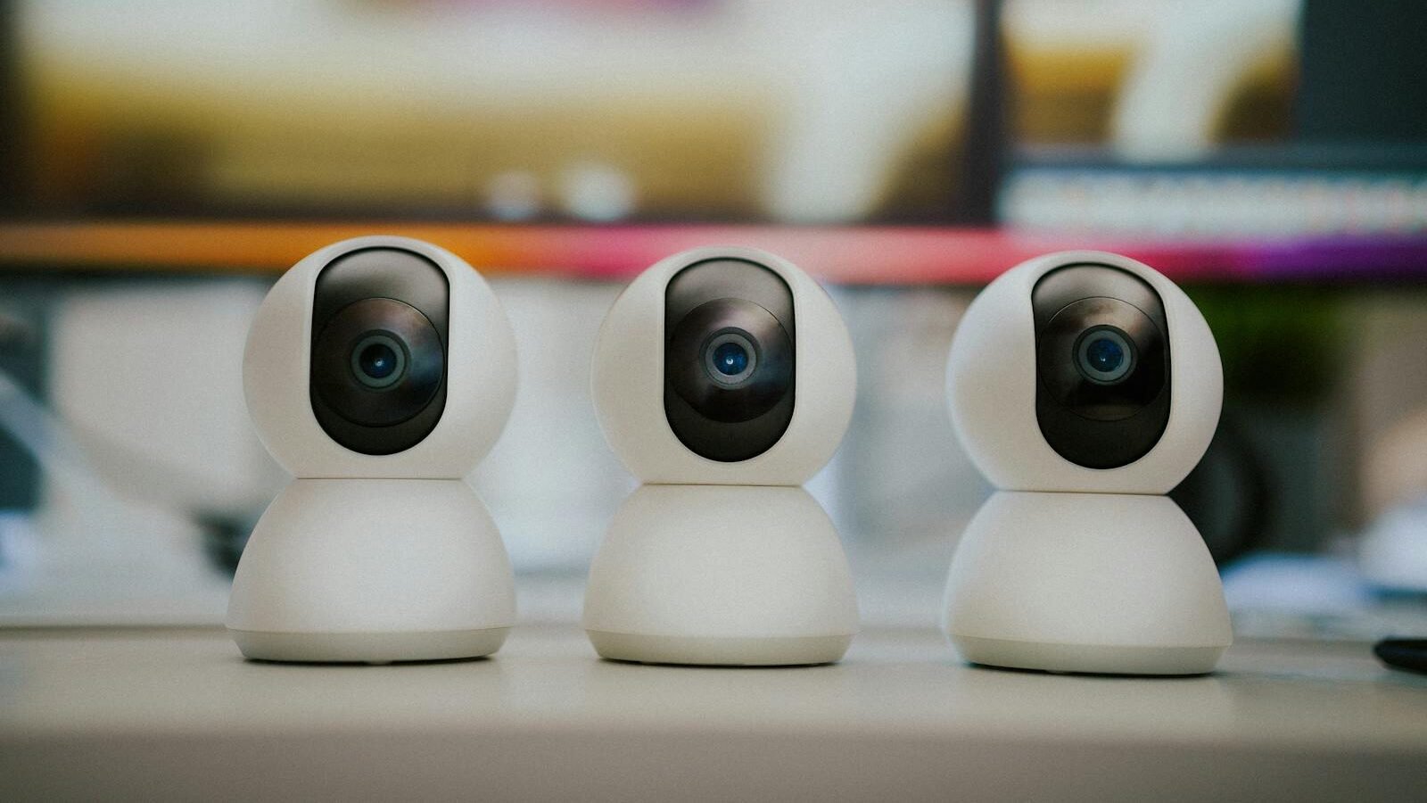 Three modern white security cameras with sleek designs placed on a desk, symbolizing advancements in monitoring and protection technology, with a blurred background.
