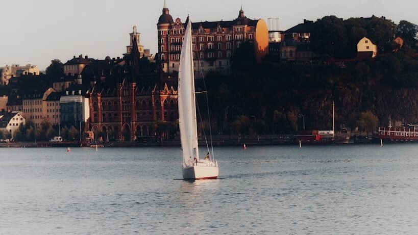 A sailboat gliding across calm waters with a backdrop of historic buildings and trees on a hillside, captured during a serene evening.