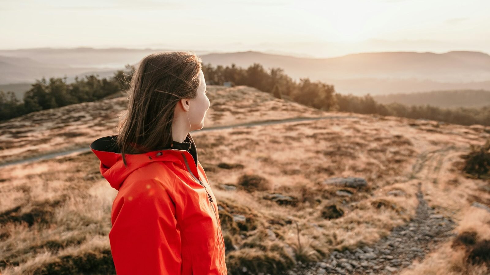 A young woman wearing an orange jacket standing on a hilltop, gazing into the distance as the sun sets over a scenic landscape.