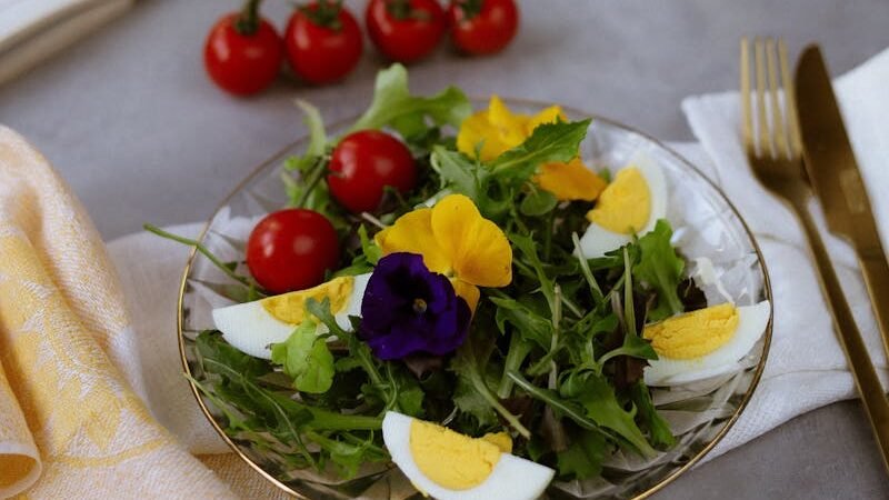 A fresh and colorful salad with leafy greens, cherry tomatoes, sliced boiled eggs, and edible flowers, served on a plate with a gold fork and knife. A bottle of olive oil, salt, a lemon, and an open book are placed on a kitchen counter in the background.