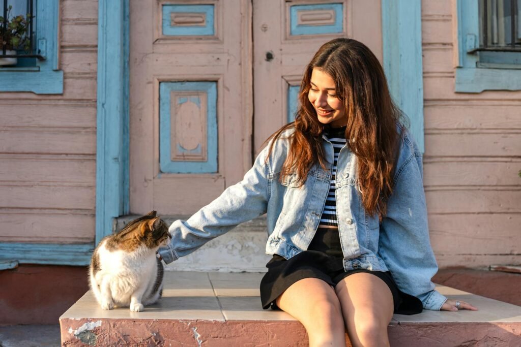 A young woman in casual attire is sitting and petting a cat on a charming rustic porch.