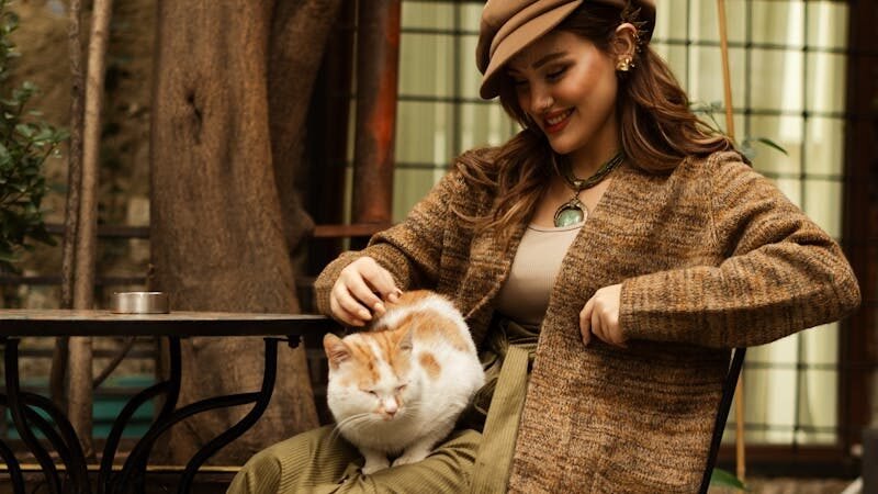 A woman in a cozy outfit sits at an outdoor café, gently petting a cat on her lap, reflecting a deep bond and understanding of feline behavior.