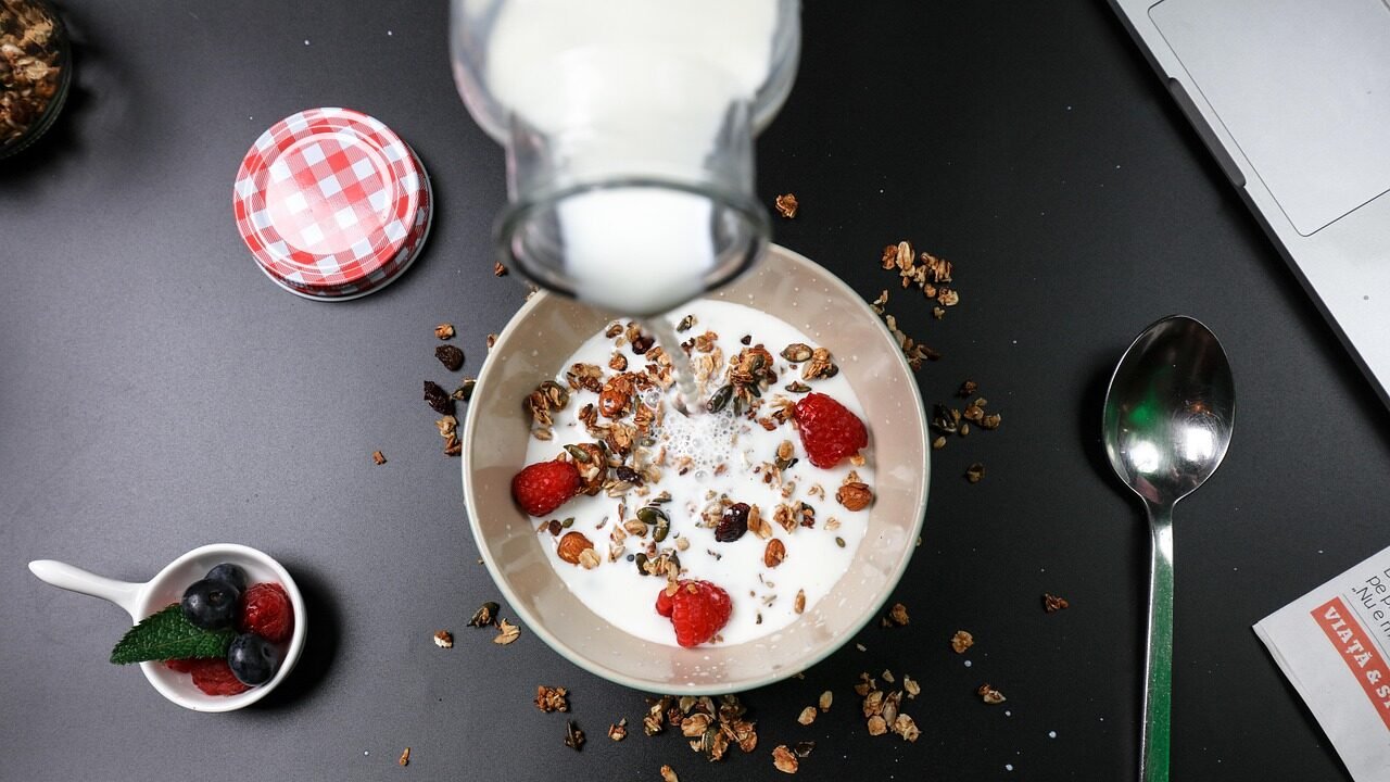 A bowl of yogurt topped with granola, raspberries, and seeds, with milk being poured over it. A small cup of mixed berries sits next to the bowl.