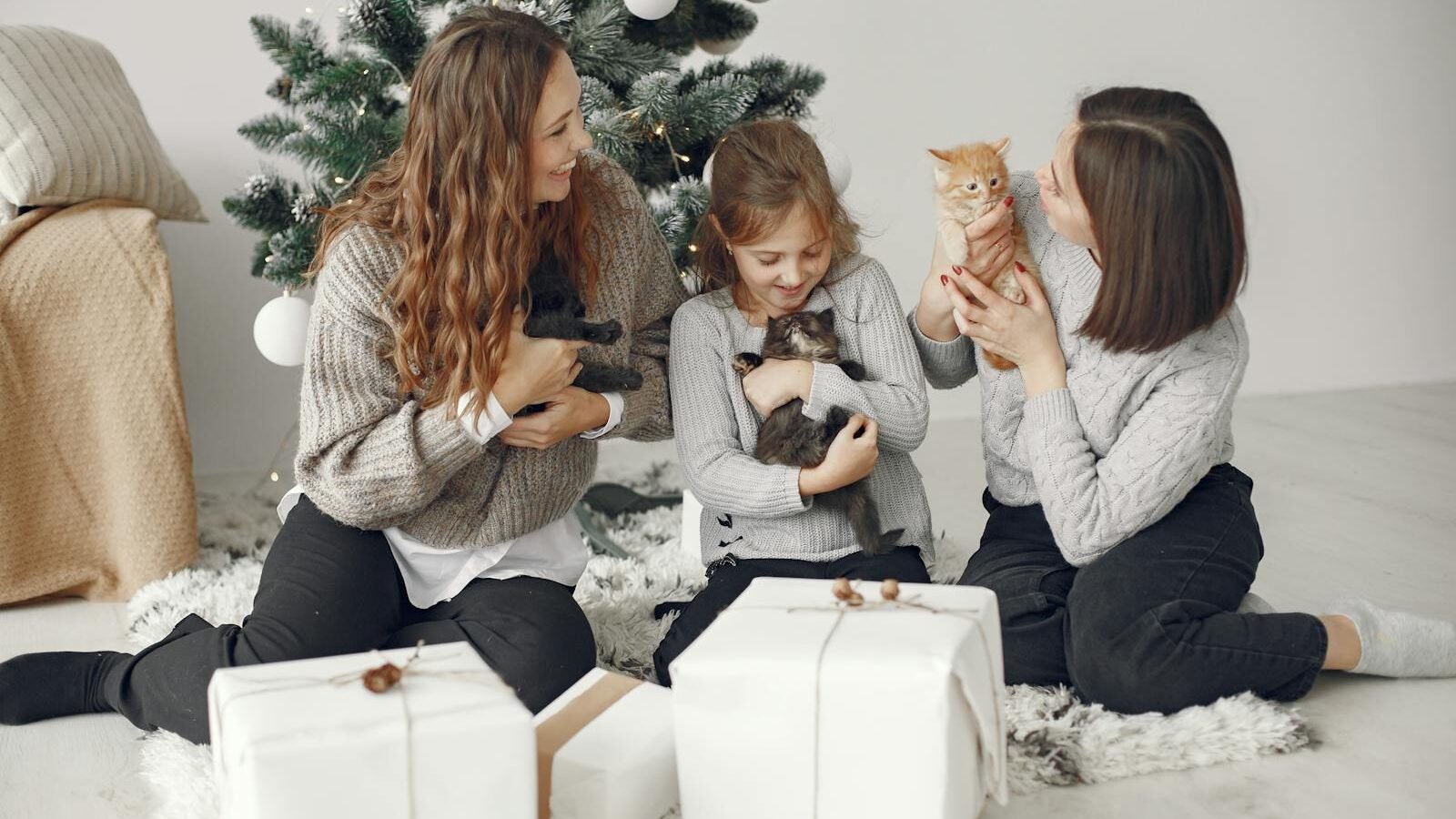 Three women and a young girl sitting around a Christmas tree with wrapped gifts, joyfully holding and playing with kittens, symbolizing the warmth and love of gifting pets during the holiday season.