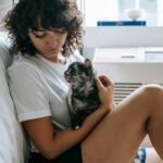 cat realationship: side view of crop young female with curly hair in casual clothes hugging and stroking adorable purebred cat while relaxing at home